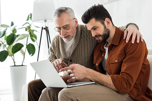 handsome bearded man pointing with finger at laptop while sitting with senior father