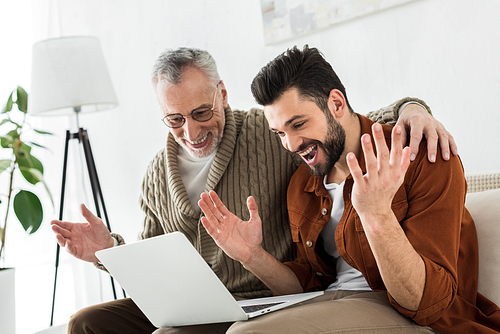 excited bearded man gesturing while sitting with senior father and looking at laptop