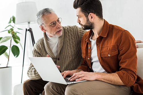 senior father looking at bearded son sitting with laptop