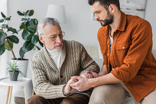 upset senior father in glasses holding hands with handsome son at home