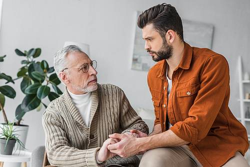 sad senior father in glasses holding hands with handsome son at home