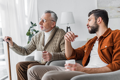 senior father in glasses sitting with handsome man and holding cup