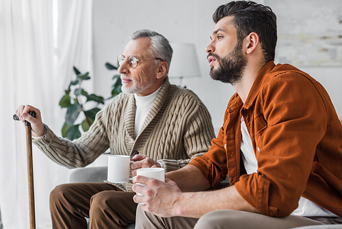 retired father in glasses sitting with handsome man and holding cup
