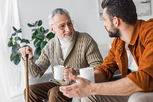 happy retired father in glasses looking at handsome son holding cup