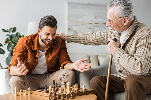 retired father in glasses putting hand on shoulder of happy son while playing chess