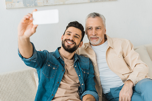 cheerful bearded man taking selfie with senior father at home