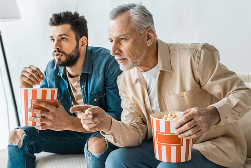 senior father pointing with finger near handsome son with popcorn bucket