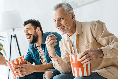 happy senior father sitting with cheerful son and holding popcorn bucket