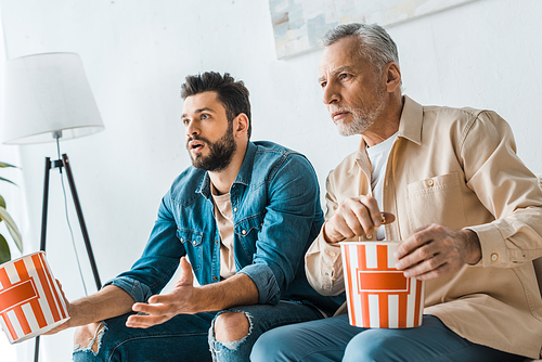 surprised senior father sitting with handsome son and holding popcorn bucket while watching tv