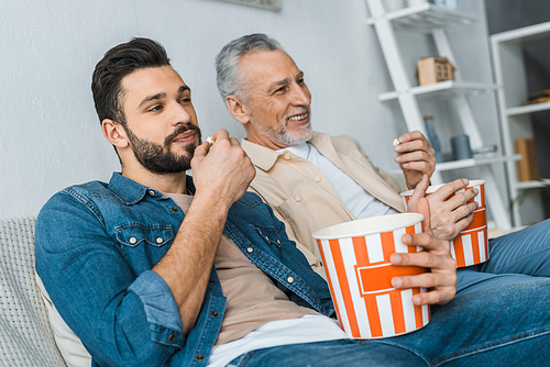 happy senior father sitting with handsome son and holding popcorn bucket while watching tv