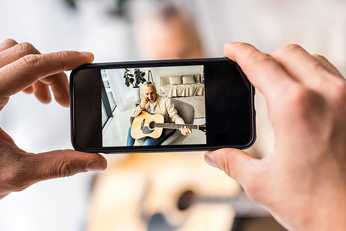 cropped view of man taking photo of senior father holding acoustic guitar at home