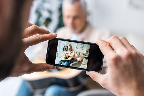 cropped view of man taking photo of retired father playing acoustic guitar at home