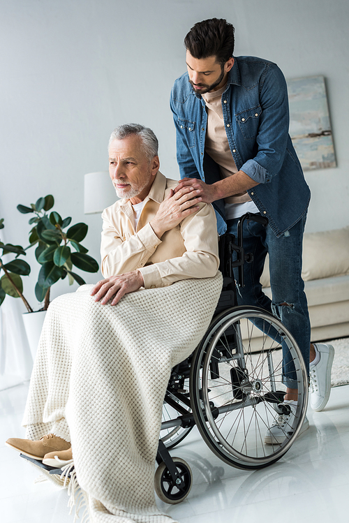 handsome bearded son holding hands with disabled senior father in wheelchair at home