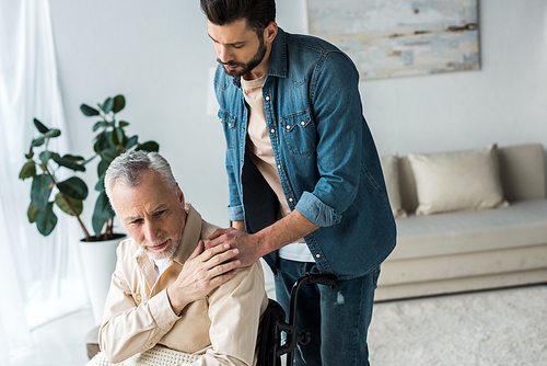 caring handsome son holding hands with disabled senior father in wheelchair at home