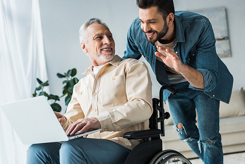 cheerful disabled retired man sitting in wheelchair and looking at handsome son waving hand while having video call on laptop