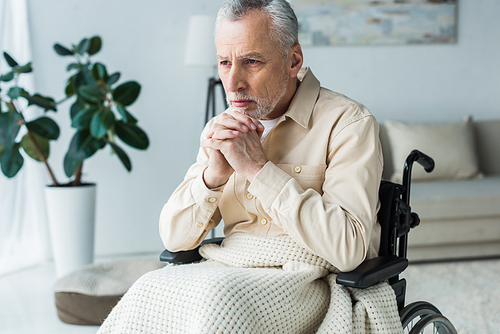 pensive disabled retired man sitting in wheelchair with clenched hands at home