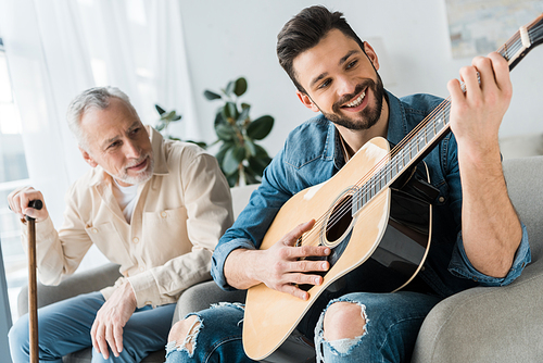 cheerful bearded man playing acoustic guitar near senior father at home
