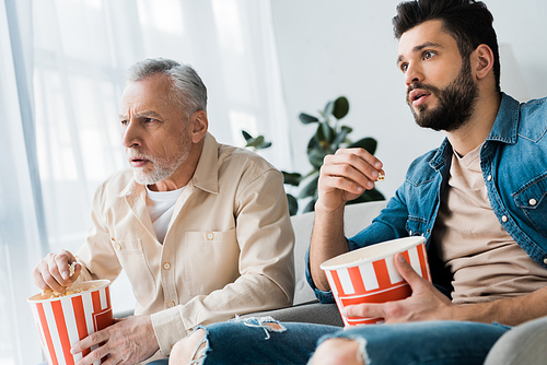 shocked senior father sitting with son and holding popcorn bucket while watching tv