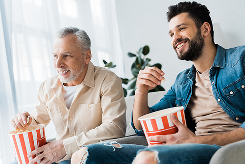 happy retired father sitting with son and holding popcorn bucket while watching tv