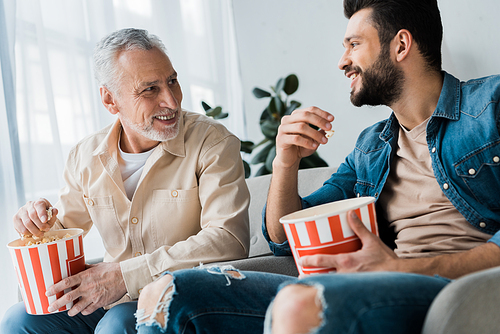 happy retired father looking at son and holding popcorn bucket at home