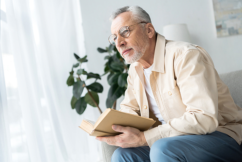 senior man in glasses holding book while sitting at home