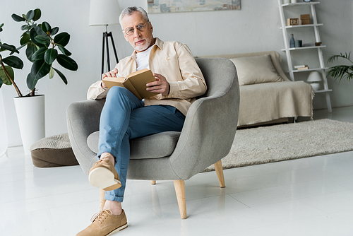 pensive retired man in glasses holding book while sitting in armchair at home