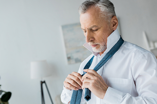 elegant senior man tying tie while standing at home