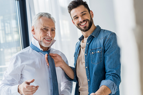 happy son helping cheerful retired father tying blue tie at home