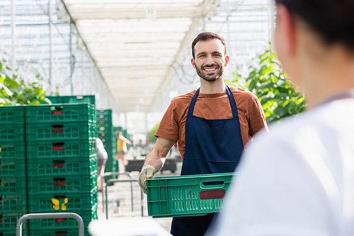 cheerful farmer holding plastic box in greenhouse near colleague on blurred foreground