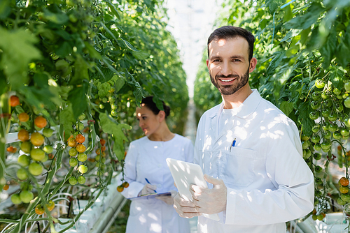 smiling quality inspector with digital tablet near african american colleague working on blurred background