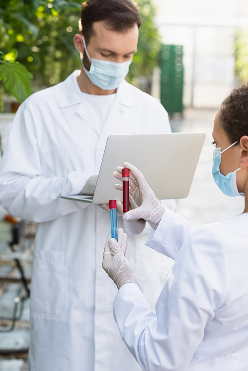 african american quality inspector in medical mask holding test tubes near colleague with laptop