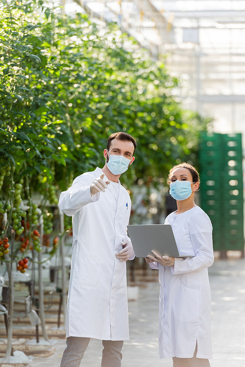 quality inspector in medical mask pointing with finger near african american colleague with laptop