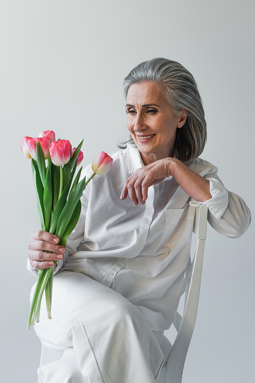 Grey haired woman in white shirt looking at flowers on chair isolated on grey