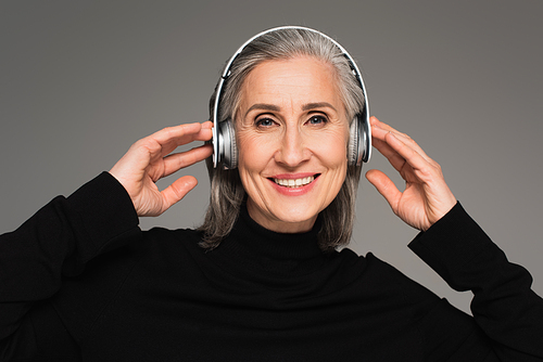 Mature woman in wireless headphones smiling at camera isolated on grey