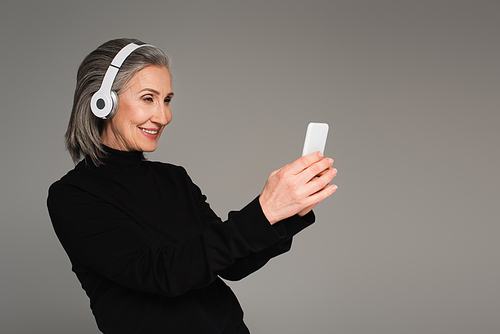 Smiling mature woman in headphones using smartphone isolated on grey