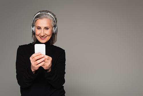 Middle aged woman in headphones using smartphone isolated on grey