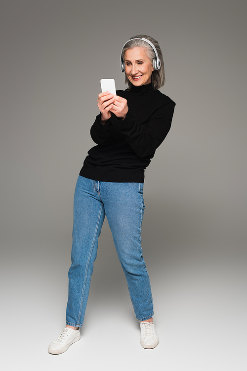 Smiling woman in jeans and jumper using smartphone and headphones on grey background