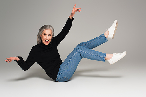 Positive mature woman in jeans sitting on grey background