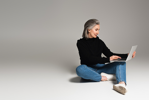Woman in turtleneck and jeans using laptop on grey background