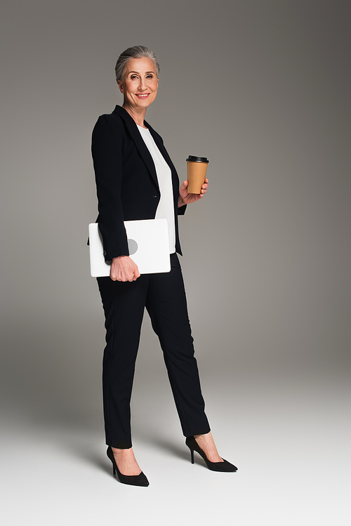 Smiling businesswoman holding takeaway drink and laptop on grey background