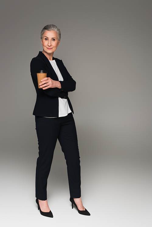 Mature businesswoman in formal wear holding paper cup on grey background