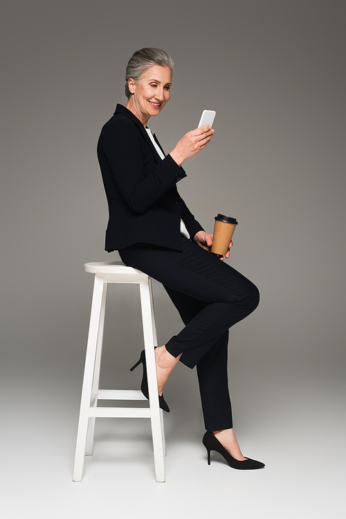 Smiling businesswoman holding paper cup and using cellphone on chair on grey background