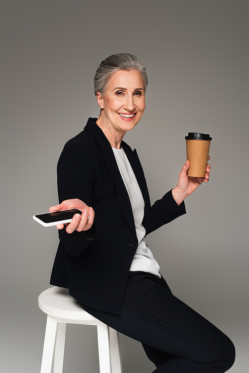 Positive mature businesswoman holding smartphone and paper cup on chair on grey background