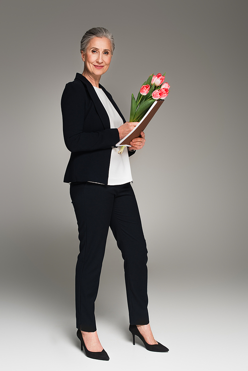 Smiling businesswoman holding paper folder and tulips on grey background