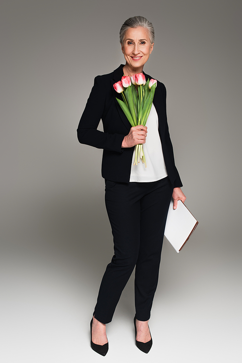 Positive businesswoman with paper folder holding tulips on grey background