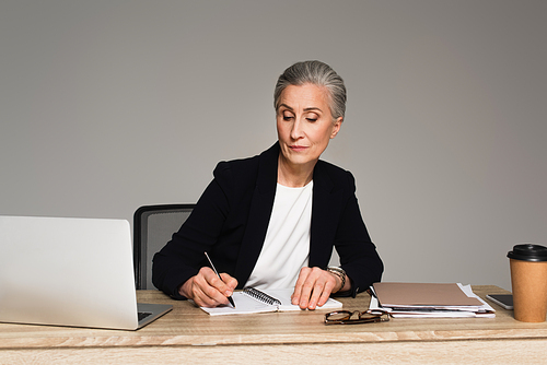 Businesswoman writing on notebook near gadgets and papers isolated on grey