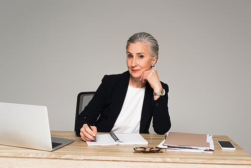 Mature businesswoman writing on notebook near papers and gadgets isolated on grey