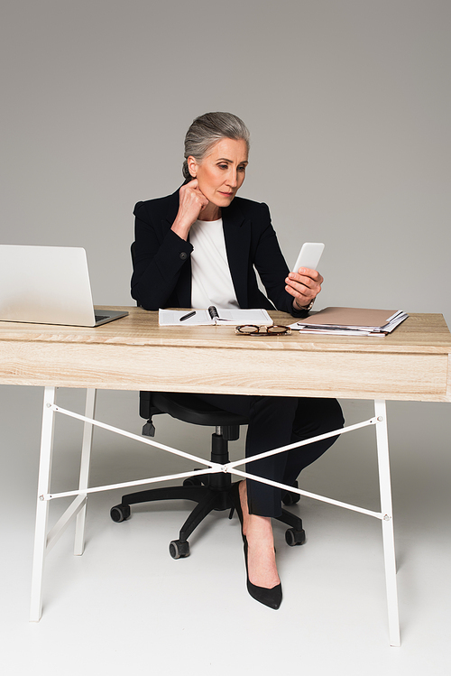 Mature businesswoman using smartphone near laptop and documents on grey background