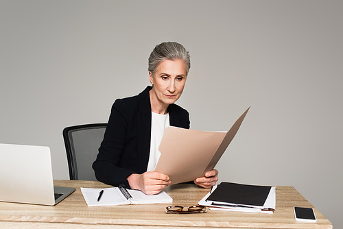 Businesswoman looking at documents near devices on table isolated on grey
