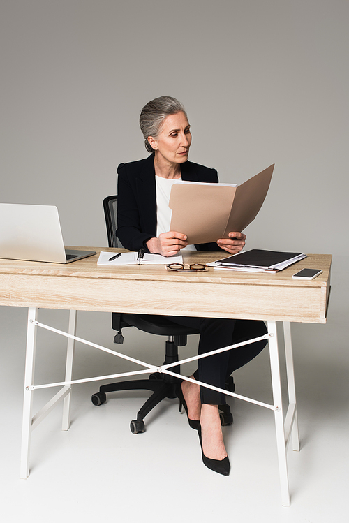 Mature businesswoman looking at documents near gadgets and eyeglasses on table on grey background